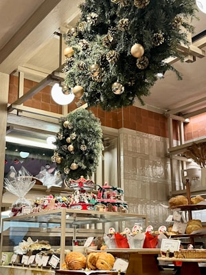 A bakery display filled with various pastries and baked goods, decorated with festive wreaths adorned with gold ornaments and pinecones. The shelves hold an assortment of bread and wrapped gift baskets, while a carousel centerpiece adds a whimsical touch.