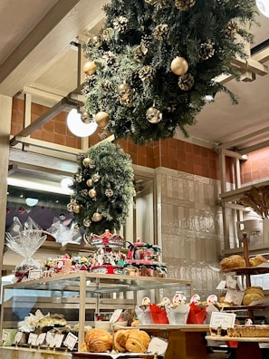 A bakery display filled with various pastries and baked goods, decorated with festive wreaths adorned with gold ornaments and pinecones. The shelves hold an assortment of bread and wrapped gift baskets, while a carousel centerpiece adds a whimsical touch.