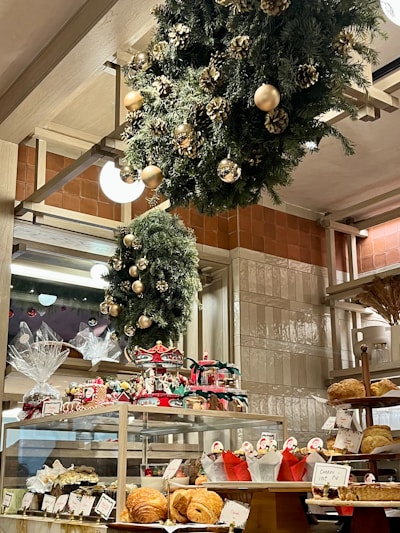 A bakery display filled with various pastries and baked goods, decorated with festive wreaths adorned with gold ornaments and pinecones. The shelves hold an assortment of bread and wrapped gift baskets, while a carousel centerpiece adds a whimsical touch.