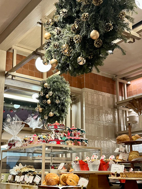 A bakery display filled with various pastries and baked goods, decorated with festive wreaths adorned with gold ornaments and pinecones. The shelves hold an assortment of bread and wrapped gift baskets, while a carousel centerpiece adds a whimsical touch.