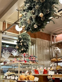 A bakery display filled with various pastries and baked goods, decorated with festive wreaths adorned with gold ornaments and pinecones. The shelves hold an assortment of bread and wrapped gift baskets, while a carousel centerpiece adds a whimsical touch.