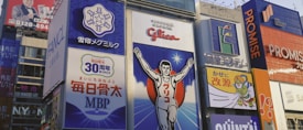 Brightly colored billboards with various advertisements line a bustling street. A prominent image of a running athlete with arms raised is featured at the center, surrounded by Japanese text and logos. Other signs include cartoon figures and corporate branding, creating a vibrant and energetic urban scene.