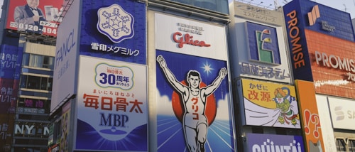 Brightly colored billboards with various advertisements line a bustling street. A prominent image of a running athlete with arms raised is featured at the center, surrounded by Japanese text and logos. Other signs include cartoon figures and corporate branding, creating a vibrant and energetic urban scene.