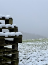 A stack of wooden pallets partially covered with snow stands in the foreground against a backdrop of a snowy field. The sky is overcast and grey, creating a cold and wintry atmosphere. The snow lightly covers the grass and pallets, suggesting a recent snowfall.