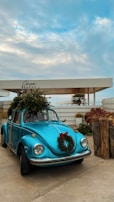 A vintage blue Volkswagen Beetle is parked with a festive wreath on its front and a Christmas tree secured on its roof. The sky is partly cloudy and there's a sign that reads 'Carvo Bohème.' The surroundings include some plants and rustic wooden stumps.