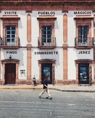 A historic building with light-colored walls and dark window frames is adorned with signs containing the words 'Visite Pueblos Mágicos', 'Pinos', 'Sombrerete', and 'Jerez'. At street level, a person in casual attire walks across the cobblestone street.