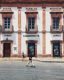 A historic building with light-colored walls and dark window frames is adorned with signs containing the words 'Visite Pueblos M&aacute;gicos', 'Pinos', 'Sombrerete', and 'Jerez'. At street level, a person in casual attire walks across the cobblestone street.