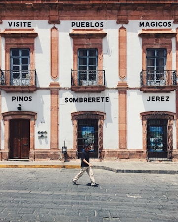 A historic building with light-colored walls and dark window frames is adorned with signs containing the words 'Visite Pueblos M&aacute;gicos', 'Pinos', 'Sombrerete', and 'Jerez'. At street level, a person in casual attire walks across the cobblestone street.