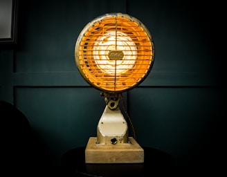 A vintage industrial-style lamp with a glowing, warm orange light is mounted on a wooden base. The lamp features a metal cage around the bulb and is set against a dark, moody background.