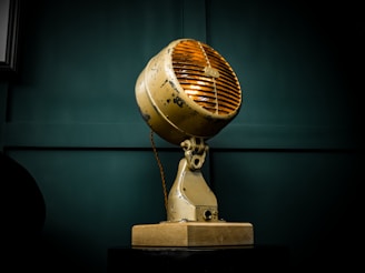 A vintage industrial lamp with a yellowish, weathered exterior. It has a round shape with a grated front cover, mounted on a sturdy base. The background features a dark green paneled wall, enhancing the lamp’s antique aesthetic.
