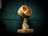 A vintage industrial lamp with a yellowish, weathered exterior. It has a round shape with a grated front cover, mounted on a sturdy base. The background features a dark green paneled wall, enhancing the lamp’s antique aesthetic.