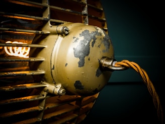 A close-up of an industrial-style light fixture with a worn, metallic surface and a visible filament glowing warmly. The frame includes a weathered protective metal grille and textured paint, giving it a vintage or rustic appearance.