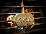 A vintage metal grille with the letters 'G.E.C.' embossed in the center. Behind the grille, there is a warm, orange glow, possibly from a light or flame, creating a striking contrast with the metallic texture.