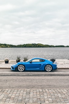 A vibrant blue sports car is parked beside a scenic lakeside view. The car is positioned on a cobblestone street with neatly lined potted plants and a wide expanse of water in the background. The sky is overcast, creating a calm and serene atmosphere.