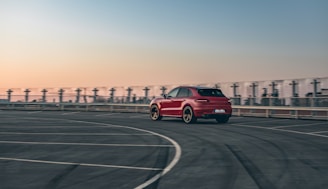 A red car is driving on an empty, spacious rooftop parking lot with marked lanes. The background features a clear sky transitioning from orange to blue hues, suggesting either sunrise or sunset. The urban skyline is faintly visible, adding to the ambient setting.