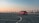 A red car is driving on an empty, spacious rooftop parking lot with marked lanes. The background features a clear sky transitioning from orange to blue hues, suggesting either sunrise or sunset. The urban skyline is faintly visible, adding to the ambient setting.