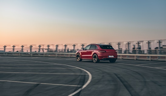 A red car is driving on an empty, spacious rooftop parking lot with marked lanes. The background features a clear sky transitioning from orange to blue hues, suggesting either sunrise or sunset. The urban skyline is faintly visible, adding to the ambient setting.