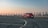 A red car is driving on an empty, spacious rooftop parking lot with marked lanes. The background features a clear sky transitioning from orange to blue hues, suggesting either sunrise or sunset. The urban skyline is faintly visible, adding to the ambient setting.