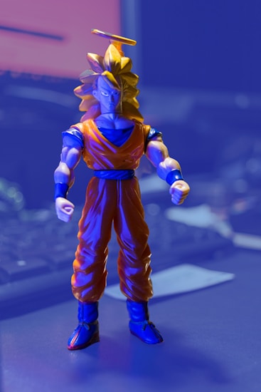 A figure of a character with spiky golden hair, an orange and blue outfit, and a golden halo above its head. The setting appears to be indoors with a blurred background that includes a keyboard and monitor, suggesting a desk environment.
