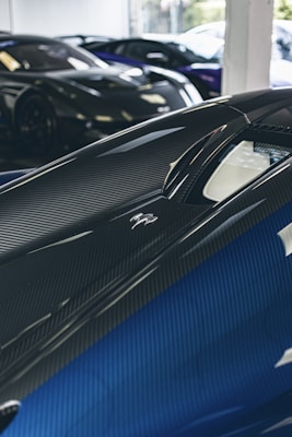 A close-up view of a sleek, modern sports car with a glossy carbon fiber finish and blue accents. The perspective highlights the curves and aerodynamics of the vehicle. In the background, other luxury cars are partially visible, indicating a high-end car showroom or garage.