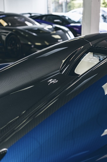 A close-up view of a sleek, modern sports car with a glossy carbon fiber finish and blue accents. The perspective highlights the curves and aerodynamics of the vehicle. In the background, other luxury cars are partially visible, indicating a high-end car showroom or garage.