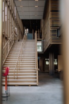 The image depicts a modern, multi-story building interior featuring a large staircase with wooden railings. The space is well-lit, with contemporary architectural elements, including a grid-like pattern on the walls and ceiling. There are two red chairs to the left side of the image.