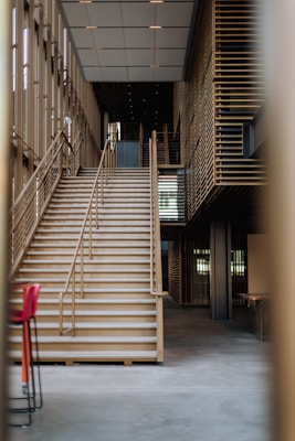 The image depicts a modern, multi-story building interior featuring a large staircase with wooden railings. The space is well-lit, with contemporary architectural elements, including a grid-like pattern on the walls and ceiling. There are two red chairs to the left side of the image.