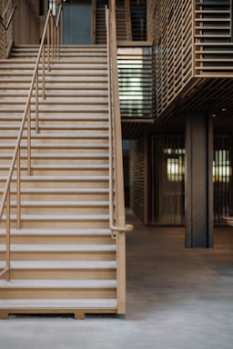 Wooden stairs with metal railings lead up to the next floor inside a modern interior space. The design features slatted wooden panels and an open area beneath the stairs, giving the structure a contemporary look.