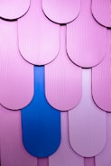 A close-up view of layered, scalloped tiles with a textured surface in shades of pink, highlighted by a single distinct blue tile, forming a pattern.