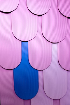 A close-up view of layered, scalloped tiles with a textured surface in shades of pink, highlighted by a single distinct blue tile, forming a pattern.
