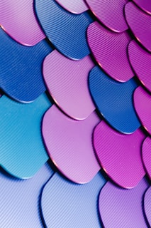 A close-up view of overlapping textured scales in vibrant colors such as purple, blue, and teal. The scales have a wavy pattern and are arranged in a repeating, harmonious sequence, creating a visually appealing pattern.