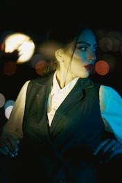 A person dressed in a dark vest and white shirt looks to the side, illuminated by a dramatic lighting setup creating a moody atmosphere. The background is blurred with bokeh lights, enhancing the cinematic effect.