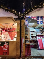 Two Christmas market stalls are side by side, adorned with festive decorations. The left stall features a person dressed in a Santa-themed sweater and hat, surrounded by holiday-themed goods and a Santa Claus poster. The right stall displays a variety of products on shelves with Canadian flags and red plaid decorations.