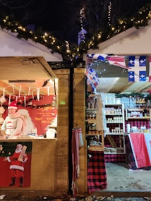 Two Christmas market stalls are side by side, adorned with festive decorations. The left stall features a person dressed in a Santa-themed sweater and hat, surrounded by holiday-themed goods and a Santa Claus poster. The right stall displays a variety of products on shelves with Canadian flags and red plaid decorations.
