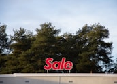 A red sign with the word 'Sale' is mounted on the roof of a building, positioned against a backdrop of tall green trees under a cloudy sky.
