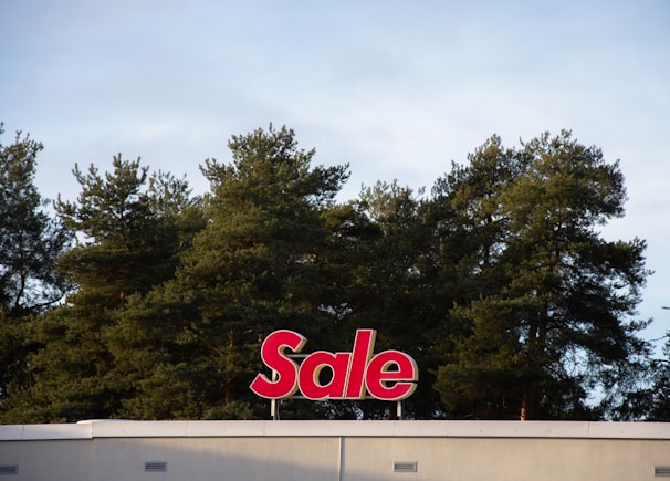 A red sign with the word 'Sale' is mounted on the roof of a building, positioned against a backdrop of tall green trees under a cloudy sky.