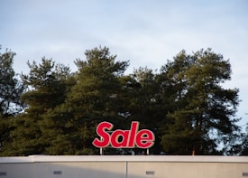 A red sign with the word 'Sale' is mounted on the roof of a building, positioned against a backdrop of tall green trees under a cloudy sky.
