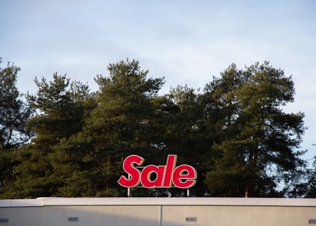 A red sign with the word 'Sale' is mounted on the roof of a building, positioned against a backdrop of tall green trees under a cloudy sky.