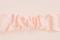 A ruffled, silky fabric in a soft peach color stretched horizontally across a neutral background. The fabric's texture creates delicate waves and folds, giving it a luxurious and smooth appearance.