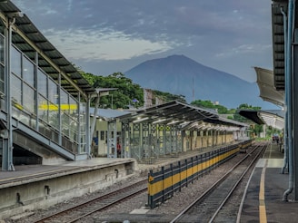 A railway station platform with multiple tracks is visible, featuring modern architecture with glass walls and metal structures. In the background, a large mountain rises, partially covered by clouds. There are a few people on the platform and lush greenery surrounding the area.