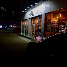 A well-lit restaurant with large glass windows, showcasing a warm and inviting interior. The exterior is lined with decorative plants and neon signs, while a dark, parked car is visible in the lower right corner. The surroundings include a dimly lit street with quiet, nighttime ambiance.