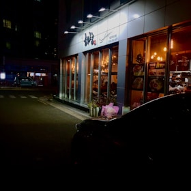 A well-lit restaurant with large glass windows, showcasing a warm and inviting interior. The exterior is lined with decorative plants and neon signs, while a dark, parked car is visible in the lower right corner. The surroundings include a dimly lit street with quiet, nighttime ambiance.
