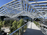 A modern structure with a complex, white steel framework forms a canopy over a landscaped area. There are lush green trees and plants along the paths, and well-designed seating areas. Large glass-walled buildings are visible in the background.