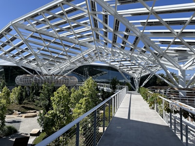 A modern structure with a complex, white steel framework forms a canopy over a landscaped area. There are lush green trees and plants along the paths, and well-designed seating areas. Large glass-walled buildings are visible in the background.