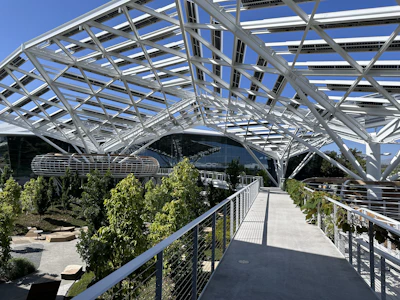 A modern structure with a complex, white steel framework forms a canopy over a landscaped area. There are lush green trees and plants along the paths, and well-designed seating areas. Large glass-walled buildings are visible in the background.