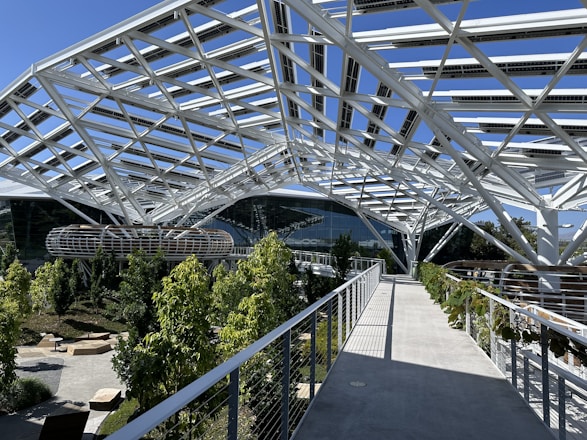 A modern structure with a complex, white steel framework forms a canopy over a landscaped area. There are lush green trees and plants along the paths, and well-designed seating areas. Large glass-walled buildings are visible in the background.