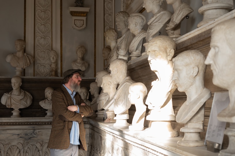 A man with a beard and a hat stands in front of a display of classical marble busts arranged in a museum-like setting. The room features ornate architectural details and is bathed in soft, warm light that highlights the textures of both the stone sculptures and the man's clothing.