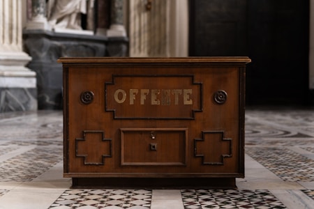 A wooden offertory box with the word 'OFFERTE' prominently displayed on its front panel. The box is situated inside a building with ornate marble flooring and tall columns visible in the background. The structure is decorated with geometric patterns and circular motifs.