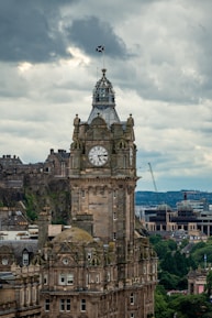 A tall clock tower with intricate architectural details stands prominently against a cloudy sky. The building features ornate stonework and a Scottish flag atop the spire. In the background, there are hills and additional historic buildings visible.