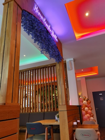 A modern interior space with wooden paneling and neon signage. The ceiling features colored lighting in purple and red tones. Balloons in gold, pink, and white are arranged against a wall. A dining area with a wooden table and cushioned seating is visible, with a plant adding greenery.